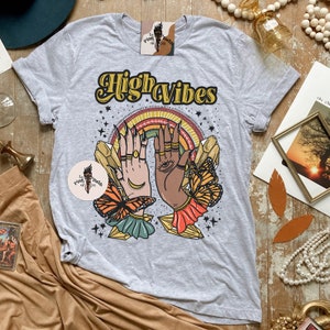 May include: A grey t-shirt with the text "High Vibes" in yellow and orange lettering. The shirt features a graphic of two hands with crystals, butterflies, and stars.