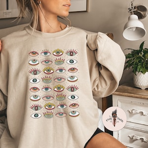 May include: A beige sweatshirt with a colorful eye pattern. The eyes are in various colors and styles, including green, blue, yellow, and pink. The sweatshirt has a crew neck and long sleeves.