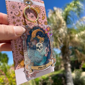 Puede incluir: Un llavero transparente con un marco dorado y una ilustraci&oacute;n colorida de una sirena y una calavera con la palabra "Pisces" en la parte inferior. El llavero est&aacute; sobre un fondo rosa con estrellas, lunas y ojos.
