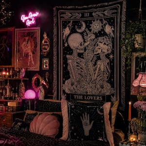 May include: A dark room with a black and white tapestry featuring skeletons and the text "THE LOVERS." The room includes a bed with decorative pillows, a neon sign that says "You Grow Girl," and various framed artworks and mirrors.