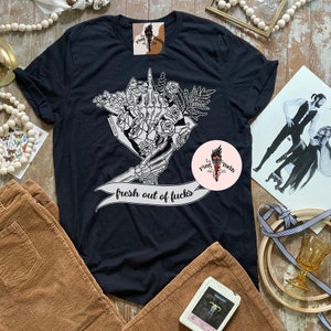 K&ouml;nnte beinhalten: Schwarzes T-Shirt mit einem Grafikdesign einer Skeletthand, die einen Blumenstrau&szlig; h&auml;lt. Der Text "fresh out of fucks" ist auf einem Banner zu sehen. Das Bild zeigt auch Cordhosen und eine gerahmte Illustration.