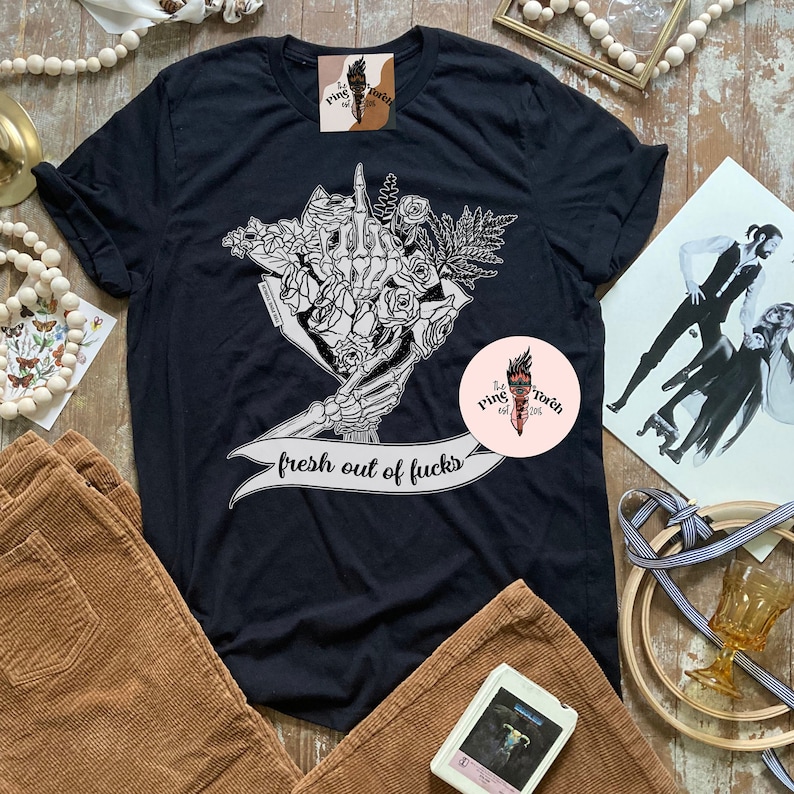 K&ouml;nnte beinhalten: Schwarzes T-Shirt mit einem Grafikdesign einer Skeletthand, die einen Blumenstrau&szlig; h&auml;lt. Der Text "fresh out of fucks" ist auf einem Banner zu sehen. Das Bild zeigt auch Cordhosen und eine gerahmte Illustration.