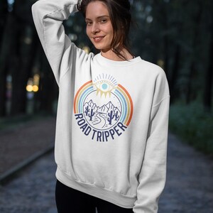 Peut inclure: Sweat-shirt blanc avec un graphique r&eacute;tro repr&eacute;sentant une cha&icirc;ne de montagnes, une route sinueuse et des cactus. Le graphique est entour&eacute; d'un arc-en-ciel et du texte "ROAD TRIPPER".