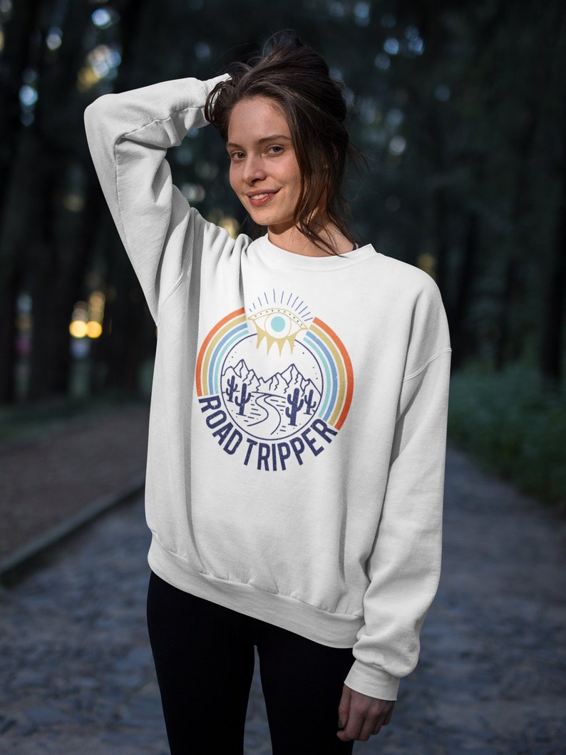 Peut inclure: Sweat-shirt blanc avec un graphique r&eacute;tro repr&eacute;sentant une cha&icirc;ne de montagnes, une route sinueuse et des cactus. Le graphique est entour&eacute; d'un arc-en-ciel et du texte "ROAD TRIPPER".