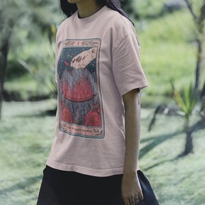 May include: A pink t-shirt with a graphic of a tarot card depicting a hand reaching down towards a burning bridge. The card is titled "The Burned Bridge".