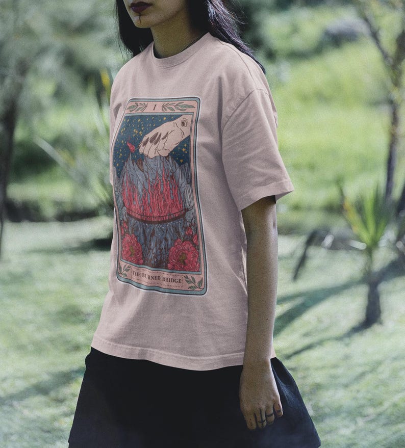 May include: A pink t-shirt with a graphic of a tarot card depicting a hand reaching down towards a burning bridge. The card is titled "The Burned Bridge".