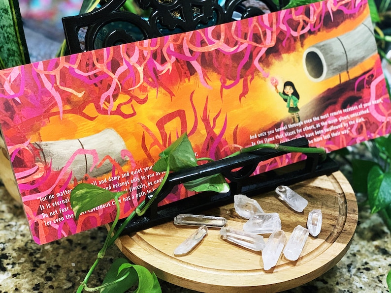 May include: A colorful bookmark with a girl holding a glowing orb in a fiery landscape. The bookmark features the text "For no matter il and damp and quiet your It is eternal and belongs only to you as fiercely, be unar And once you tumel through even the mett remote nestes of your sea Ir the flame that blares for othert, at the noen plout, unsched Do not fear for even the war cavernous Ce has a path ta he have latt their way"