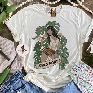 K&ouml;nnte beinhalten: Ein wei&szlig;es T-Shirt mit einem Aufdruck einer Frau in einer Dschungelkulisse. Die Frau tr&auml;gt einen Bikini mit Leopardenmuster und hat eine tropische Pflanze hinter sich. Der Text "SEND NODES" befindet sich auf einem Banner unter ihr.