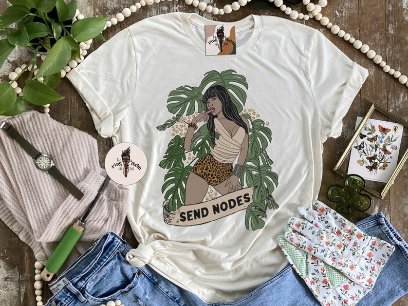 K&ouml;nnte beinhalten: Ein wei&szlig;es T-Shirt mit einem Aufdruck einer Frau in einer Dschungelkulisse. Die Frau tr&auml;gt einen Bikini mit Leopardenmuster und hat eine tropische Pflanze hinter sich. Der Text "SEND NODES" befindet sich auf einem Banner unter ihr.
