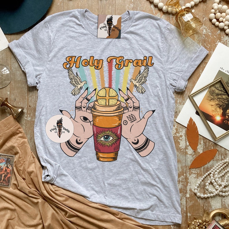 Op de afbeelding: Een grijs T-shirt met een grafisch ontwerp van twee handen die een koffiebeker vasthouden met een oog erop. De tekst "Holy Grail" is boven de beker gedrukt. Het ontwerp is in retro-stijl met regenboogstrepen en vogels.