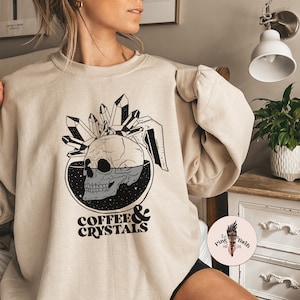 May include: A beige sweatshirt with a graphic of a skull inside a coffee pot with crystals on top. The text "COFFEE & CRYSTALS" is printed below the graphic.