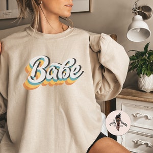 Może przedstawiać: Beżowa bluza z nadrukiem słowa "Babe" w retro tęczowej czcionce.
