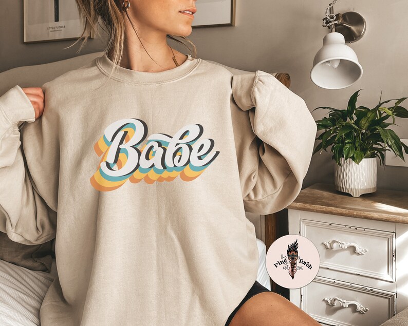 Może przedstawiać: Beżowa bluza z nadrukiem słowa "Babe" w retro tęczowej czcionce.