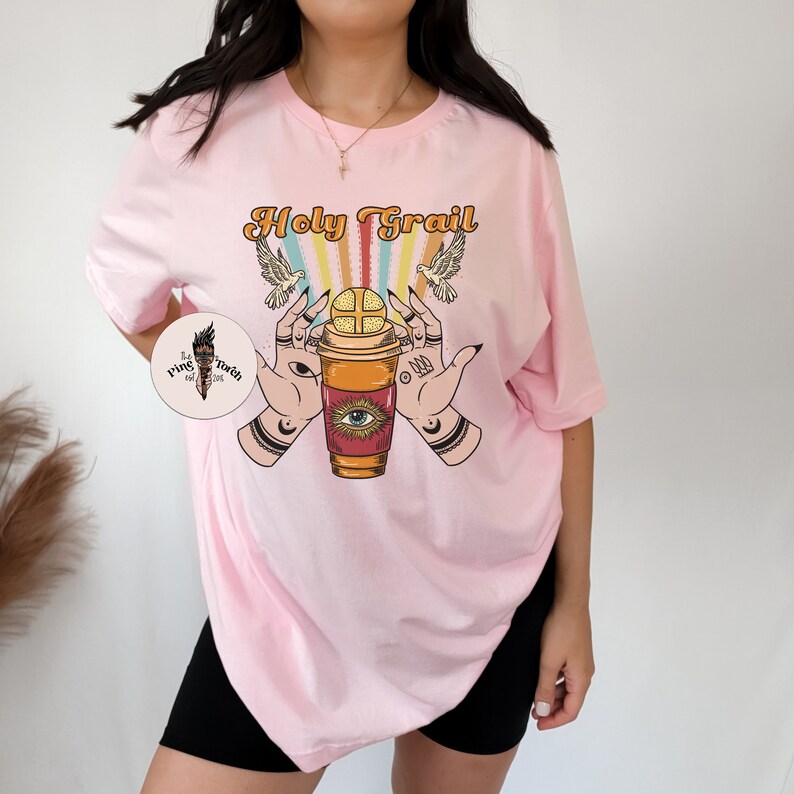 Op de afbeelding: Roze oversized t-shirt met de tekst "Holy Grail" in een retro lettertype. Het ontwerp bevat een koffiebeker met een oog en een regenboog erboven.
