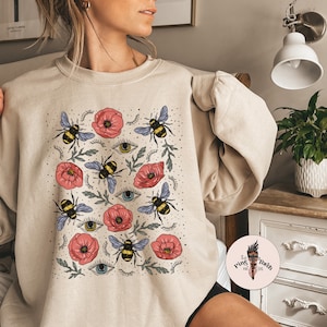 K&ouml;nnte beinhalten: Ein beiger Pullover mit einem Muster aus Bienen, Blumen und Augen. Die Blumen sind rosa und die Bienen sind gelb und schwarz.