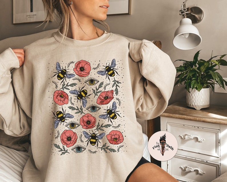 K&ouml;nnte beinhalten: Ein beiger Pullover mit einem Muster aus Bienen, Blumen und Augen. Die Blumen sind rosa und die Bienen sind gelb und schwarz.