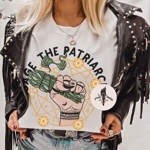 Puede incluir: Camiseta blanca con un gr&aacute;fico de una mano sosteniendo un palo de salvia. El gr&aacute;fico est&aacute; rodeado de un c&iacute;rculo con estrellas y el texto "Sage the Patriarchy".