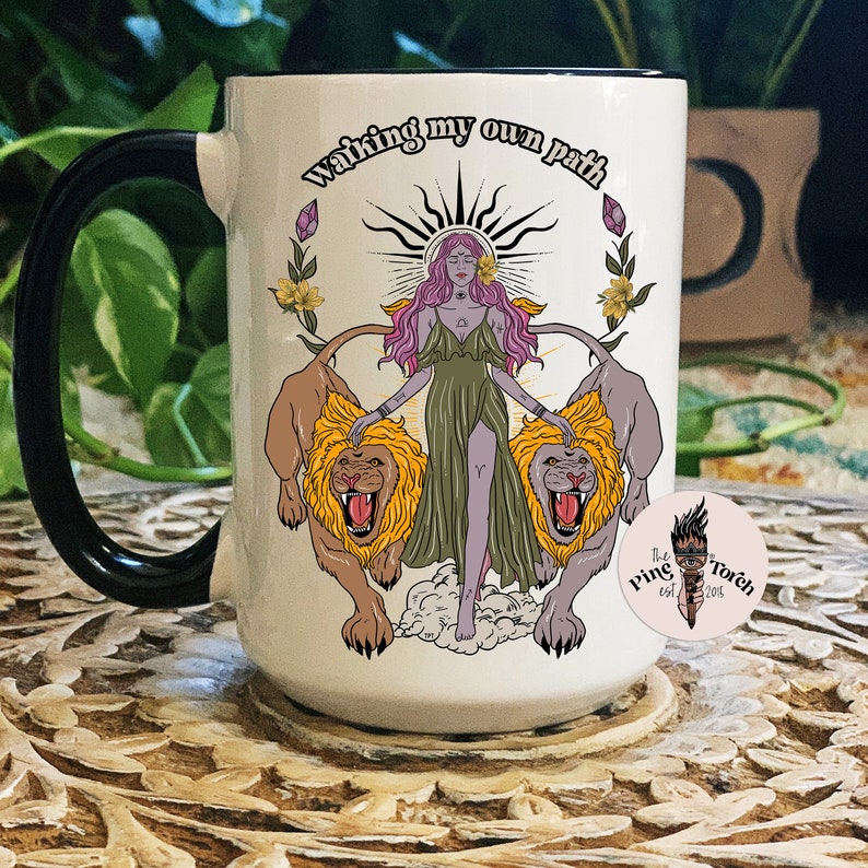 May include: White ceramic mug with a black handle. The mug features a colorful illustration of a woman standing between two lions with the text "Walking my own path".