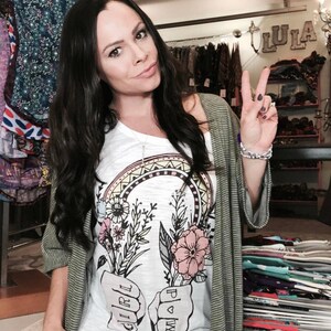 May include: A woman wearing a white t-shirt with a graphic of two fists holding flowers and the words "Girl Power" printed on them. She is also wearing a green cardigan and has her hair pulled back.