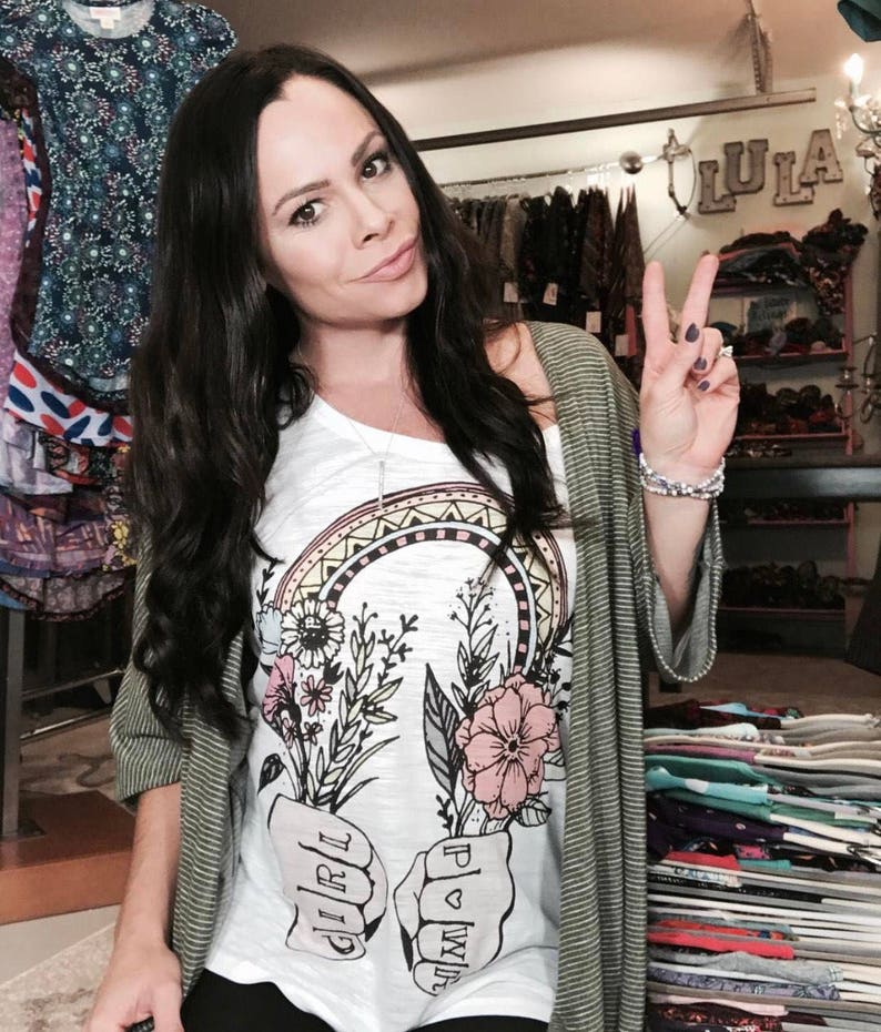May include: A woman wearing a white t-shirt with a graphic of two fists holding flowers and the words "Girl Power" printed on them. She is also wearing a green cardigan and has her hair pulled back.