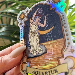 以下が含まれることがあります： ホログラフィックステッカー、水瓶座の星座デザイン。イラストは、三日月のある星空を背景に、女性が容器から水を注いでいる様子を描いています。「AQUARIUS」の文字がバナーに表示されています。