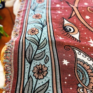 May include: A red woven tapestry with a floral border and a paisley design. The tapestry features a repeating pattern of eyes with eyelashes and stars.