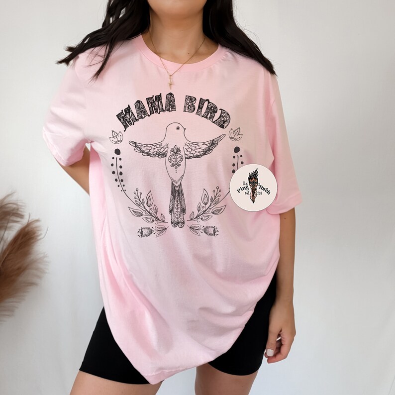 May include: Pink oversized t-shirt with a black graphic of a bird with the text "Mama Bird" and a small logo in the bottom right corner.