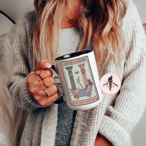 K&ouml;nnte beinhalten: Eine wei&szlig;e Keramiktasse mit schwarzem Rand und Henkel. Die Tasse zeigt eine farbenfrohe Illustration der Tarotkarte Hohepriesterin mit dem Text "The High Priestess" und "XXVI" auf der Vorderseite. Die Tasse wird von einer Person gehalten.