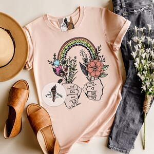 May include: A pink t-shirt with a rainbow, flowers, and the text "Girl Power" in a fist. The shirt is on a white surface with a hat, sandals, and a sticker that says "The Pine Torch".