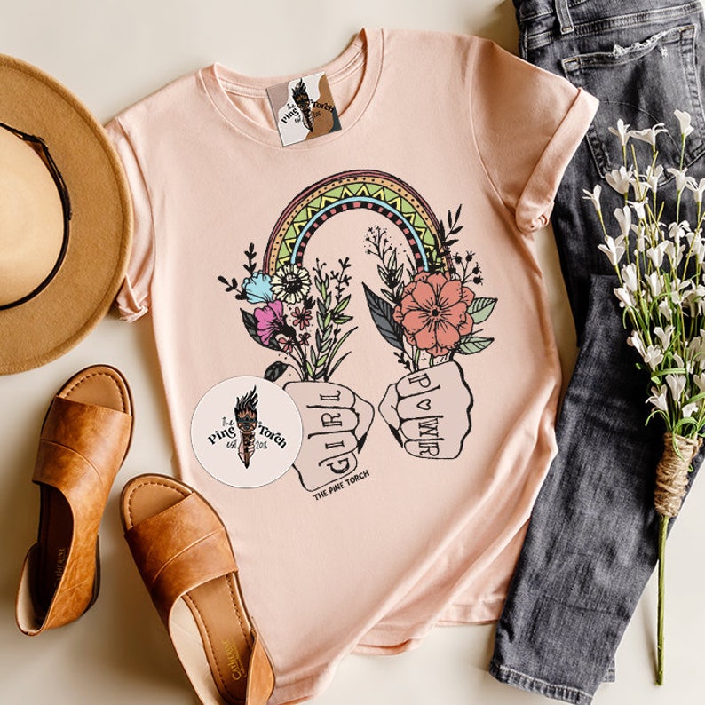 May include: A pink t-shirt with a rainbow, flowers, and the text "Girl Power" in a fist. The shirt is on a white surface with a hat, sandals, and a sticker that says "The Pine Torch".