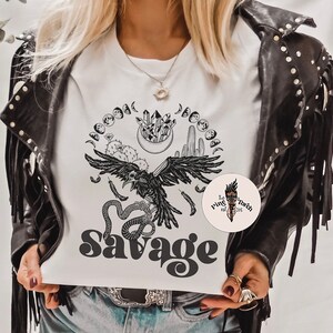 May include: White t-shirt with a black and white graphic design featuring a raven, snake, cactus, crystals, and moons. The text "Savage" is printed in black below the graphic. The t-shirt is being worn by a person with a black leather jacket.