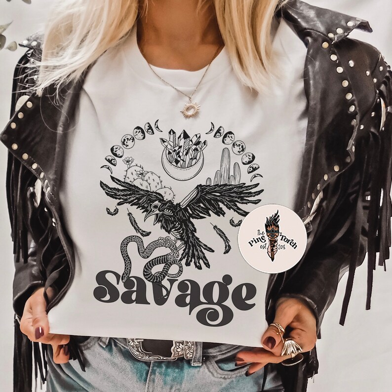 May include: White t-shirt with a black and white graphic design featuring a raven, snake, cactus, crystals, and moons. The text "Savage" is printed in black below the graphic. The t-shirt is being worn by a person with a black leather jacket.