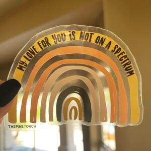May include: A clear sticker featuring a rainbow design and the text "My love for you is not on a spectrum". The rainbow is composed of various shades of brown, orange, and yellow. The sticker is set against a white backdrop.