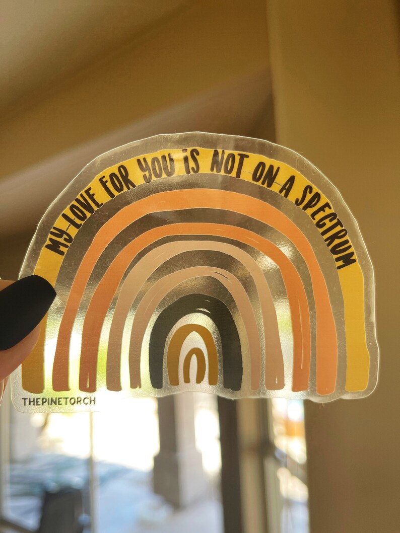 May include: A clear sticker featuring a rainbow design and the text "My love for you is not on a spectrum". The rainbow is composed of various shades of brown, orange, and yellow. The sticker is set against a white backdrop.
