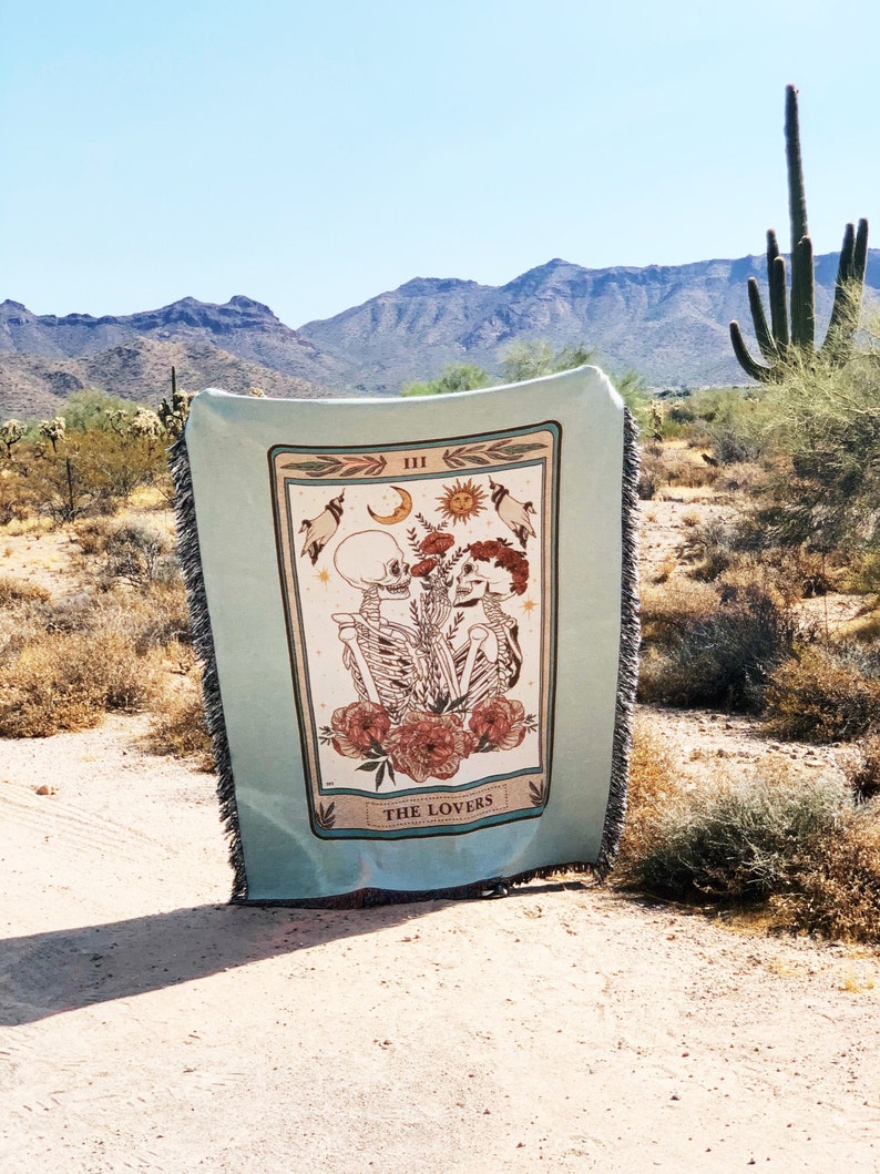 May include: A light blue woven throw blanket with a tarot card design featuring skeletons, flowers, and the words "THE LOVERS." The blanket has a fringed edge and is displayed outdoors with a desert landscape background.