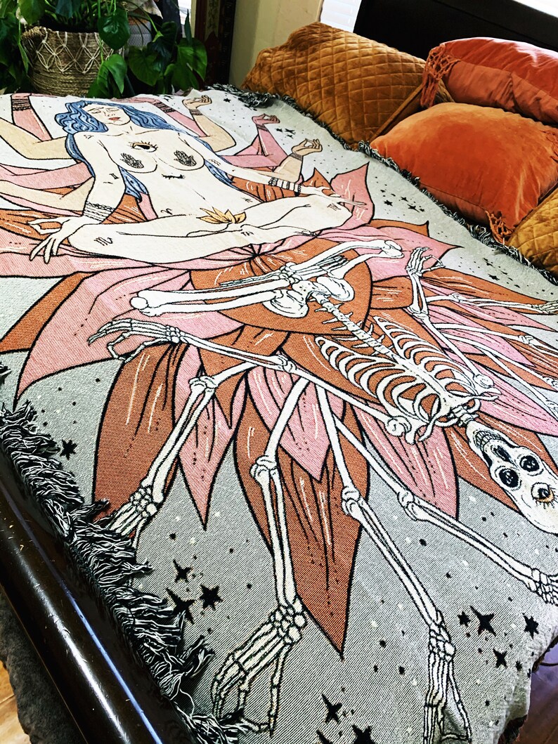 May include: A woven tapestry throw blanket with a gray background featuring a pink lotus flower with a skeleton figure and a woman with multiple arms sitting on the flower. The woman has blue hair and a third eye. The background is a dark gray with white stars.