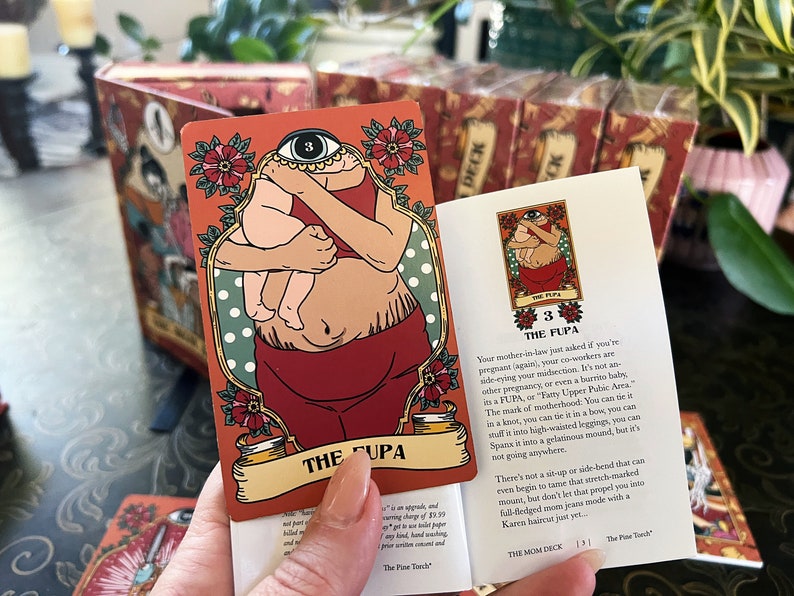 May include: A red and green illustrated card with a woman holding a baby. The card is from the "The Mom Deck" and is titled "The FUPA". The card number is 3.