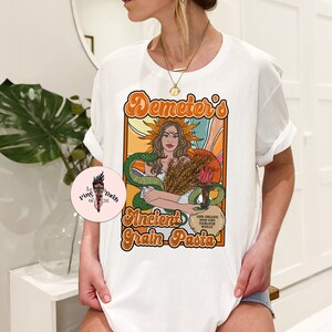 May include: White t-shirt with a graphic design featuring a woman with wheat and a snake. The text on the shirt reads "Demeter's Ancient Grain Pasta".