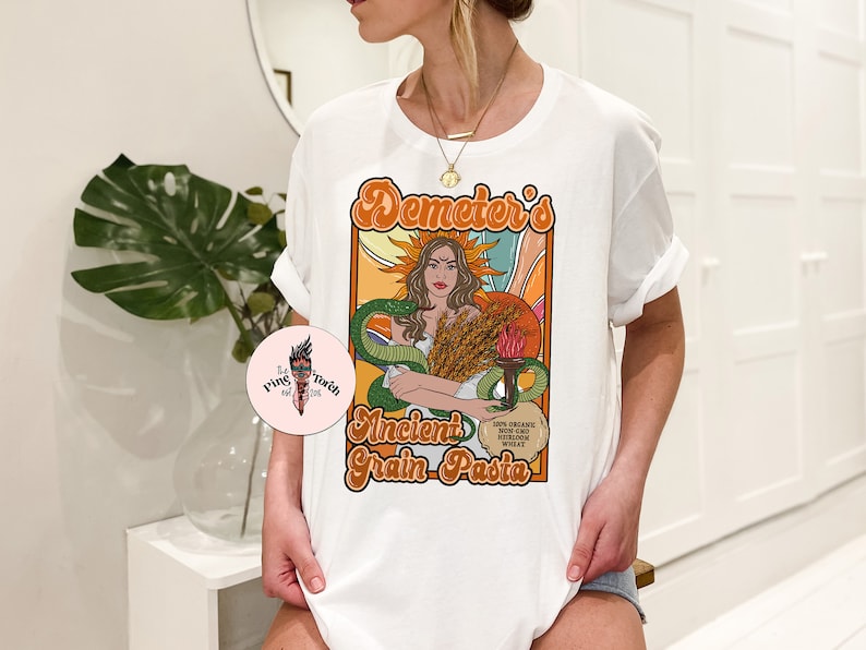 May include: White t-shirt with a graphic design featuring a woman with wheat and a snake. The text on the shirt reads "Demeter's Ancient Grain Pasta".