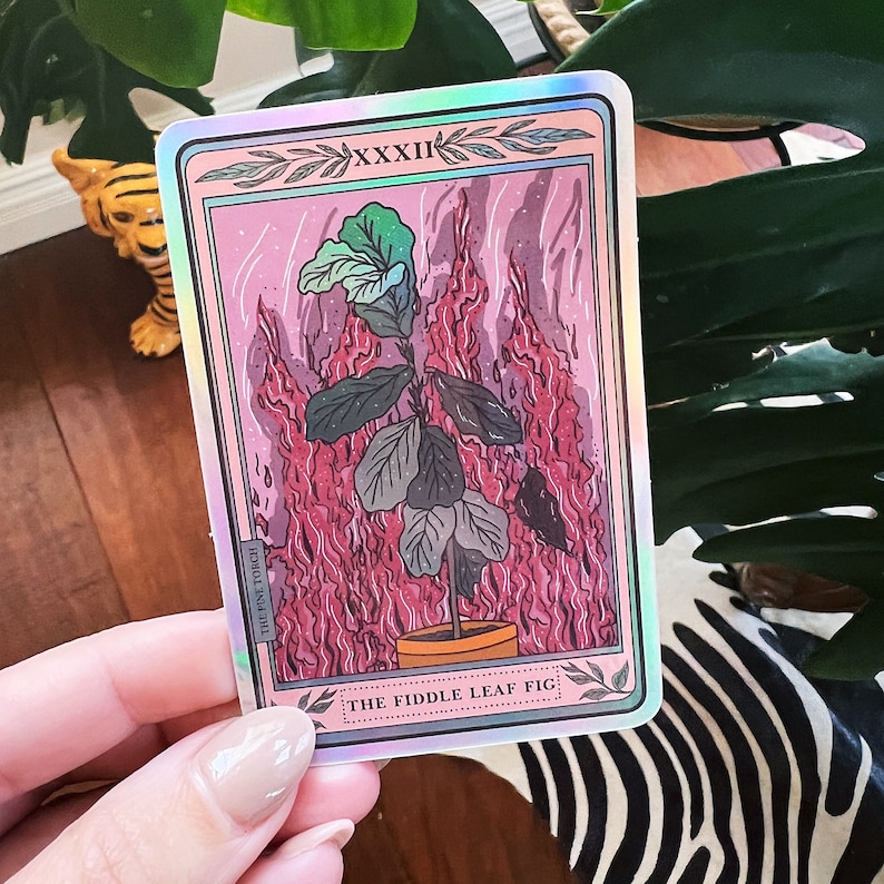 K&ouml;nnte beinhalten: Eine holografische Tarotkarte mit rosa Hintergrund und einer Zeichnung einer Geigenfeige in einem Terrakotta-Topf. Die Karte ist von Flammen umgeben und tr&auml;gt die Nummer XXXII sowie den Text "THE FIDDLE LEAF FIG".