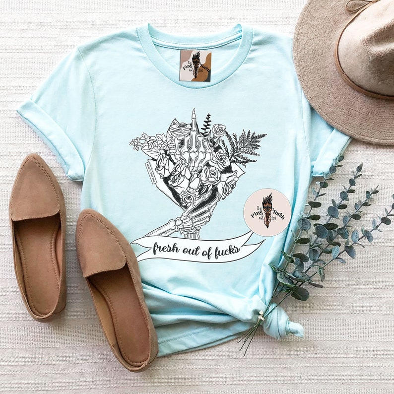K&ouml;nnte beinhalten: Hellblaues T-Shirt mit einer schwarz-wei&szlig;en Grafik einer Skeletthand, die einen Blumenstrau&szlig; h&auml;lt, mit dem Text "fresh out of fucks". Das Bild zeigt auch beige Loafer und einen beigen Hut.