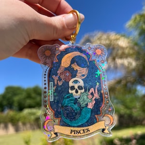 Puede incluir: Un llavero hologr&aacute;fico con una sirena y una calavera con un tercer ojo. El llavero est&aacute; en un marco dorado con la palabra "Pisces" en la parte inferior. El fondo es un cielo estrellado azul y negro.
