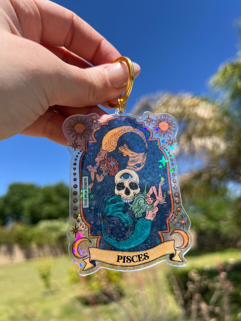 Puede incluir: Un llavero hologr&aacute;fico con una sirena y una calavera con un tercer ojo. El llavero est&aacute; en un marco dorado con la palabra "Pisces" en la parte inferior. El fondo es un cielo estrellado azul y negro.