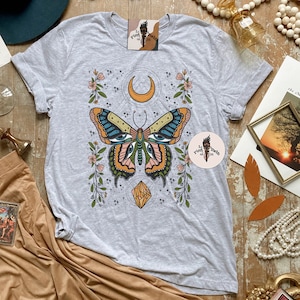 May include: A grey t-shirt with a butterfly design. The butterfly has large eyes and is surrounded by flowers and stars. The text "Pink Toech of Girl" is on the shirt.