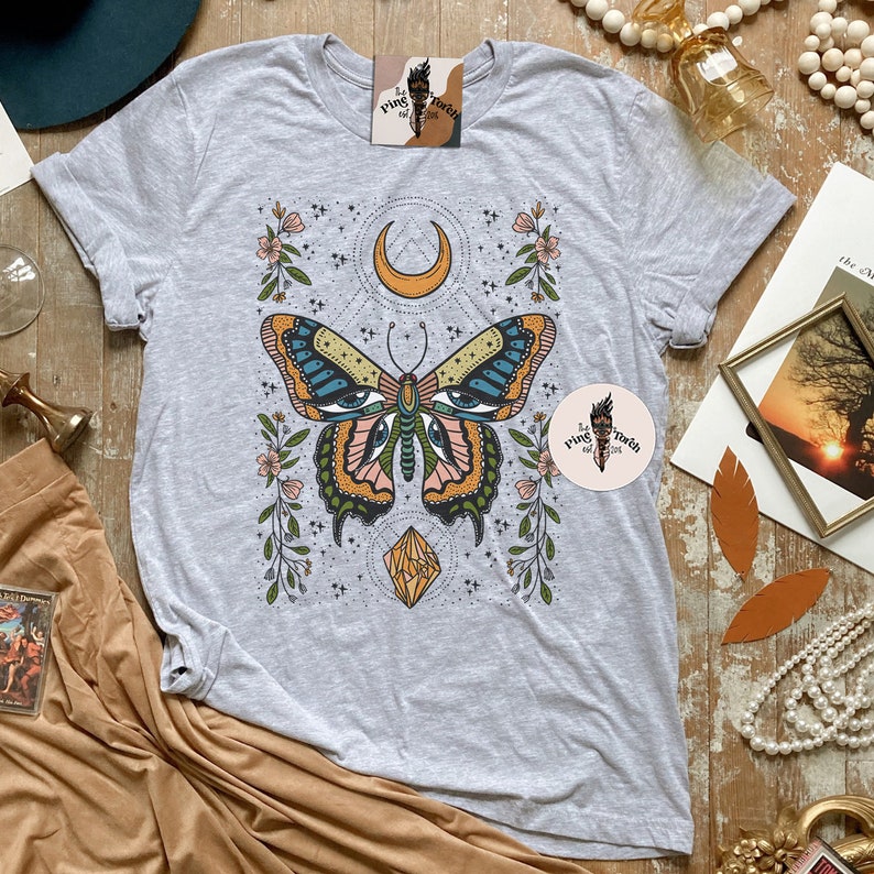 May include: A grey t-shirt with a butterfly design. The butterfly has large eyes and is surrounded by flowers and stars. The text "Pink Toech of Girl" is on the shirt.