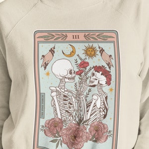 May include: A light beige sweatshirt with a graphic print of a tarot card. The card is titled "The Lovers" and features two skeletons holding flowers. The card is surrounded by a pink and blue border.