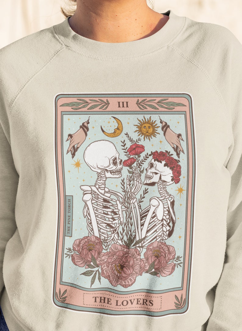 May include: A light beige sweatshirt with a graphic print of a tarot card. The card is titled "The Lovers" and features two skeletons holding flowers. The card is surrounded by a pink and blue border.