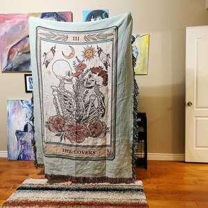 May include: A light green woven throw blanket featuring a tarot card design. The card depicts two skeletons embracing, with floral and celestial elements. The text "THE LOVERS" is at the bottom of the card.