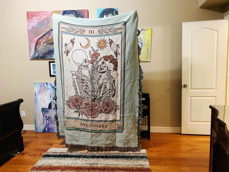 May include: A light green woven throw blanket featuring a tarot card design. The card depicts two skeletons embracing, with floral and celestial elements. The text "THE LOVERS" is at the bottom of the card.