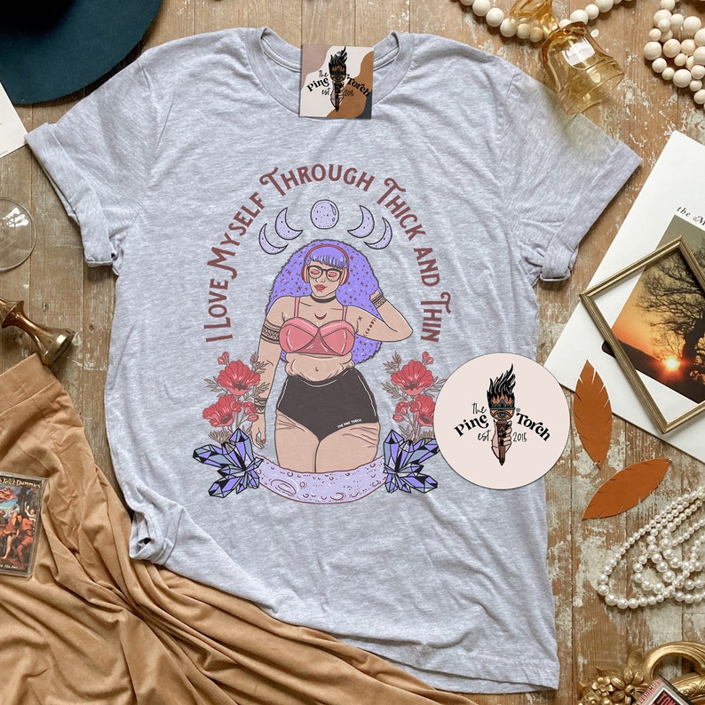 Puede incluir: Una camiseta gris con un gr&aacute;fico de una mujer con cabello morado que lleva un sujetador rosa y pantalones cortos negros. El gr&aacute;fico est&aacute; rodeado de flores, cristales y lunas. El texto "I love myself through thick and thin" est&aacute; escrito alrededor del gr&aacute;fico. El texto "The Pink Torch est. 2015" est&aacute; escrito en un c&iacute;rculo en la esquina inferior derecha.